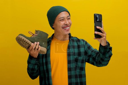 Homme asiatique dans un bonnet et des vêtements décontractés montre avec enthousiasme ses nouvelles chaussures dans un selfie. Je viens d'acheter en ligne ! Isolé sur un fond jaune vif.