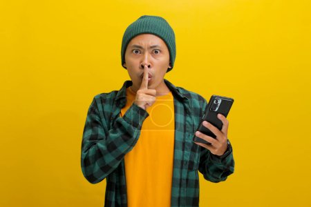 A young Asian man, dressed casually in a beanie hat and casual clothes, is gesturing for silence by placing his fingers on his lips, signaling to keep a secret while holding a phone, yellow background