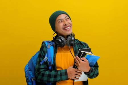 Selbstbewusster asiatischer Student in Mütze und lässiger Kleidung, mit Rucksack und Kopfhörer, einen Stapel Bücher in der Hand. Vereinzelt auf gelbem Hintergrund. Akademischer Erfolg, Vorbereitung oder die Freude am Lernen.