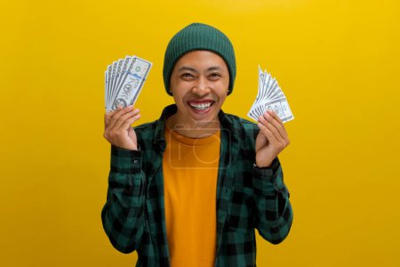 Homme asiatique excité dans un bonnet et des vêtements décontractés tient des billets de banque dans sa main. Isolé sur un fond jaune vif. Parfait pour illustrer les concepts de gain financier, d'excitation et de richesse.