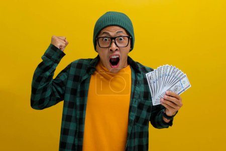 Excited Asian man wearing a beanie hat and casual clothes raising his fist making a YES gesture celebrating success while clutching money isolated over yellow background.