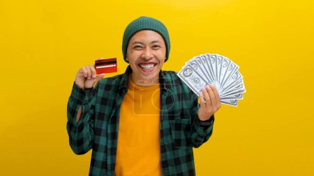 Thrilled Asian man in a beanie and casual clothes holds up a credit card and a stack of banknotes, Isolated on a yellow background. Financial success, shopping sprees, and exciting purchases concept.