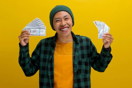 Homme asiatique excité dans un bonnet et des vêtements décontractés tient des billets de banque dans sa main. Isolé sur un fond jaune vif. Parfait pour illustrer les concepts de gain financier, d'excitation et de richesse.