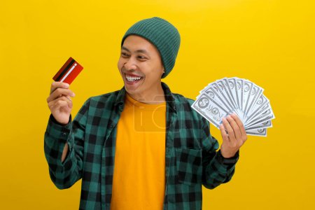 Aufgeregter asiatischer Mann in Mütze und lässiger Kleidung hält eine Kreditkarte und einen Stapel Banknoten in die Höhe, isoliert auf gelbem Hintergrund. Finanzielle Erfolge, Einkaufsbummel und spannendes Kaufkonzept.