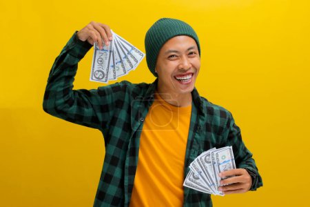 Excited Asian man in a beanie and casual clothes holds banknotes in his hand. Isolated on a bright yellow background. Perfect for illustrating concepts of financial gain, excitement, and wealth.