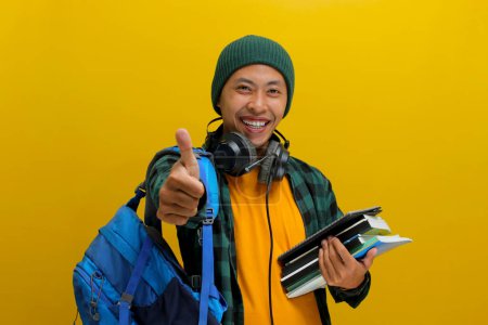 Confident Asian student in beanie and casual clothes, carrying backpack and headphones, Holding a stack of books. Isolated on yellow background. Academic success, preparation, or the joy of learning.