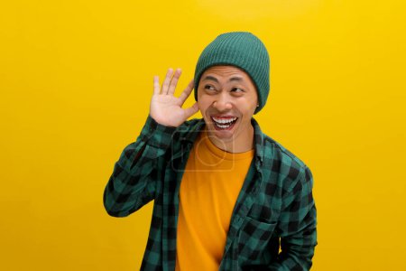 Foto de Joven hombre asiático, vestido con un sombrero de gorro y camisa casual, se ríe mientras escucha atentamente, la mano tapó su oreja, posiblemente atrapando el viento de rumores o chismes mientras está de pie sobre el fondo amarillo - Imagen libre de derechos