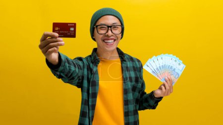 Thrilled Asian man in a beanie and casual clothes holds up a credit card and a stack of banknotes, Isolated on a yellow background. Financial success, shopping sprees, and exciting purchases concept.