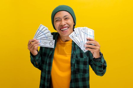 Excited Asian man in a beanie and casual clothes holds banknotes in his hand. Isolated on a bright yellow background. Perfect for illustrating concepts of financial gain, excitement, and wealth.