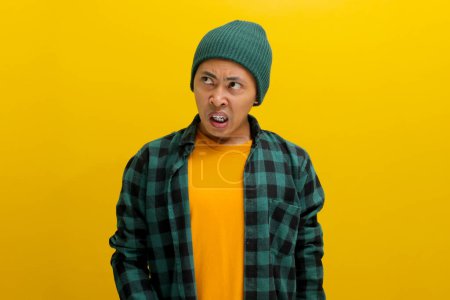 Displeased young Asian man, dressed in a beanie hat and casual shirt, is clearly expressing his disgust at something awful while standing against yellow background.