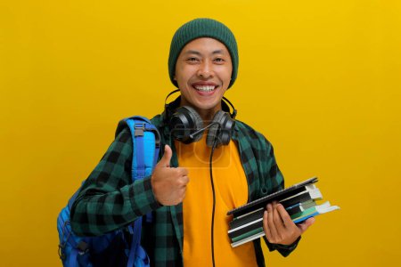 Estudiante asiática confiada en gorro y ropa casual, llevando mochila y auriculares, sosteniendo una pila de libros. Aislado sobre fondo amarillo. Éxito académico, preparación o la alegría de aprender.