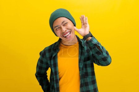 Foto de Joven hombre asiático, vestido con un sombrero de gorro y camisa casual, se ríe mientras escucha atentamente, la mano tapó su oreja, posiblemente atrapando el viento de rumores o chismes mientras está de pie sobre el fondo amarillo - Imagen libre de derechos