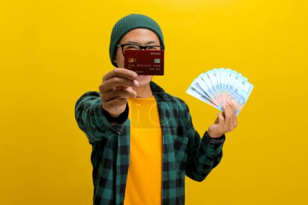Emocionado hombre asiático en un gorro y ropa casual sostiene una tarjeta de crédito y una pila de billetes, aislado sobre un fondo amarillo. Éxito financiero, compras y emocionante concepto de compras.