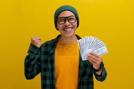 Excited Asian man wearing a beanie hat and casual clothes raising his fist making a YES gesture celebrating success while clutching money isolated over yellow background.