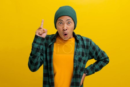 Aufgeregte junge Asiaten, mit Beanie-Hut und lässigem Hemd bekleidet, haben eine Idee, heben den Finger in einem Eureka-Zeichen und lächeln erstaunt, während sie vor gelbem Hintergrund stehen.