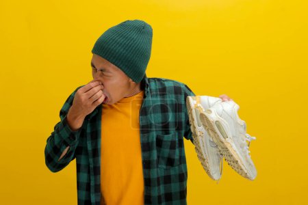 Asiatischer Mann in Mütze und kariertem Hemd grimassiert, während er seine Nase zukneift und wahrscheinlich auf einen starken Geruch reagiert. Vereinzelt auf gelbem Hintergrund. Unangenehme Gerüche, Ekel und Humor