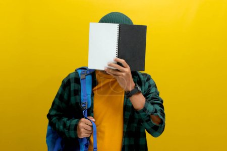 A young Asian man, dressed in a beanie hat and casual shirt and carrying a backpack, is covering his face with a book while standing against a yellow background.