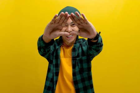 Téléchargez les photos : Jeune homme asiatique, vêtu d'une chemise décontractée et coiffé d'un bonnet, crée une forme de triangle avec ses mains, symbolisant le concept de protection de l'environnement, de recyclage et de réutilisation - en image libre de droit