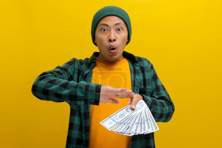 Happy Asian man in a beanie throws banknotes into the air, a joyous expression on his face. Isolated on a yellow background. Financial freedom, extravagant spending, or celebrating success concept