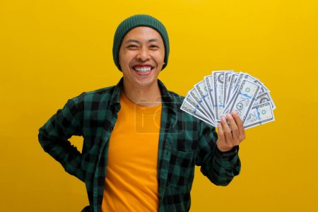 Emocionado hombre asiático en un gorro y ropa casual sostiene billetes en su mano. Aislado sobre un fondo amarillo brillante. Perfecto para ilustrar conceptos de ganancia financiera, emoción y riqueza.