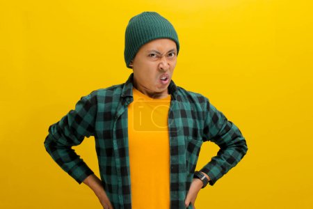 Unzufriedener junger asiatischer Mann, mit Mütze und lässigem Hemd bekleidet, drückt seinen Abscheu vor etwas Schrecklichem deutlich aus, während er vor gelbem Hintergrund steht.