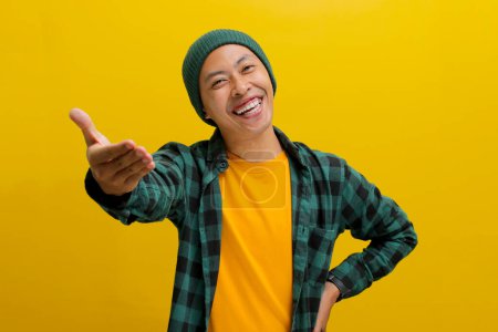 Freundlicher junger asiatischer Mann, mit Mütze und lässigem Hemd bekleidet, macht eine Handgeste, die eine Einladung signalisiert oder Hilfe anbietet, mit ausgestreckter Hand, während er vor gelbem Hintergrund steht.