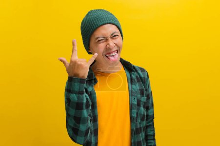 Entusiasta hombre asiático con gorro y ropa casual lanza un letrero de roca, sonriendo emocionado a la cámara con la lengua fuera. Aislado sobre un fondo amarillo brillante. Perfecto para ilustrar conceptos de conciertos, fans de la música y emoción.