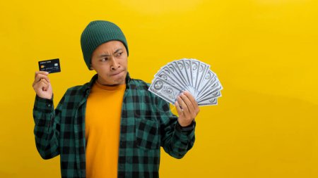Besinnlicher asiatischer Mann in Mütze und lässiger Kleidung begutachtet Kreditkarte und Bargeld und überlegt einen Einkauf. Vereinzelt auf gelbem Hintergrund. Finanzentscheidungen, Budgetierung und durchdachte Ausgaben