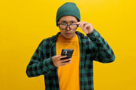 Furieux homme asiatique dans un bonnet et chemise à carreaux ajuste ses lunettes, regardant directement la caméra tout en tenant un téléphone. Isolé sur fond jaune. Frustration, colère et enjeux technologiques.