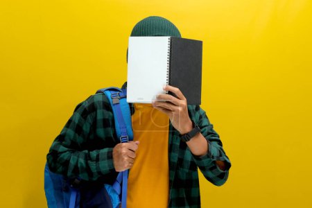 A young Asian man, dressed in a beanie hat and casual shirt and carrying a backpack, is covering his face with a book while standing against a yellow background.