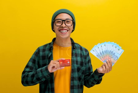 Emocionado hombre asiático en un gorro y ropa casual sostiene una tarjeta de crédito y una pila de billetes, aislado sobre un fondo amarillo. Éxito financiero, compras y emocionante concepto de compras.