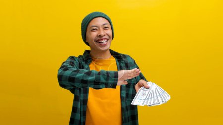 Happy Asian man in a beanie throws banknotes into the air, a joyous expression on his face. Isolated on a yellow background. Financial freedom, extravagant spending, or celebrating success concept