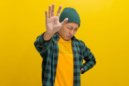 Unzufriedener junger asiatischer Mann, mit Mütze und lässigem Hemd bekleidet, drückt seinen Abscheu vor etwas Schrecklichem deutlich aus, während er vor gelbem Hintergrund steht.