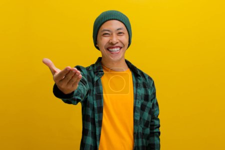 Freundlicher junger asiatischer Mann, mit Mütze und lässigem Hemd bekleidet, macht eine Handgeste, die eine Einladung signalisiert oder Hilfe anbietet, mit ausgestreckter Hand, während er vor gelbem Hintergrund steht.