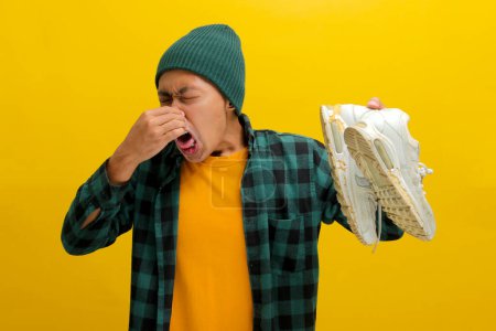 Photo for Asian man in a beanie and plaid shirt grimaces while pinching his nose shut, likely reacting to a strong odor. Isolated on a yellow background. Unpleasant smells, disgust, and humor concept - Royalty Free Image