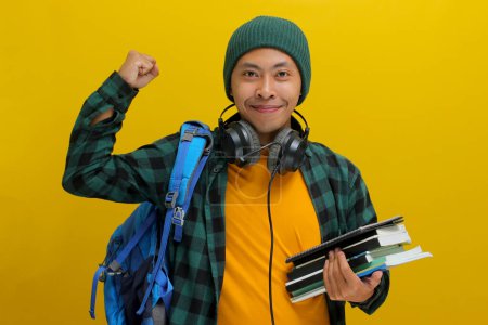 Selbstbewusster asiatischer Student in Mütze und lässiger Kleidung, mit Rucksack und Kopfhörer, einen Stapel Bücher in der Hand. Vereinzelt auf gelbem Hintergrund. Akademischer Erfolg, Vorbereitung oder die Freude am Lernen.