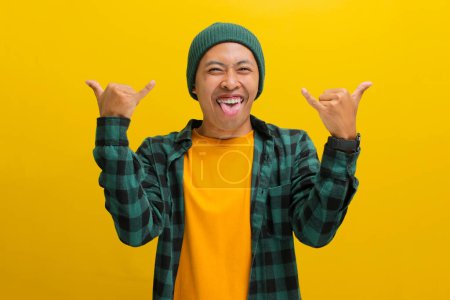 Enthusiastischer Asiate in Mütze und lässiger Kleidung wirft ein Felsschild hoch und grinst aufgeregt in die Kamera, die Zunge herausgestreckt. Vereinzelt auf leuchtend gelbem Hintergrund. Perfekt, um Konzepte von Konzerten, Musikfans und Spannung zu illustrieren.