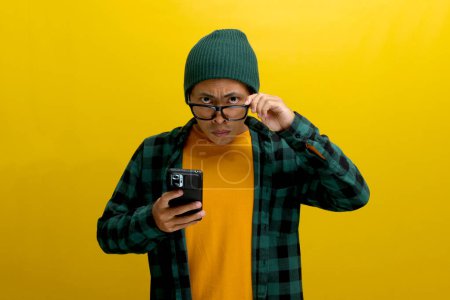 Furious Asian man in a beanie and plaid shirt adjusts his eyeglasses, glaring directly at the camera while holding a phone. Isolated on yellow background. Frustration, anger, and technological issues.