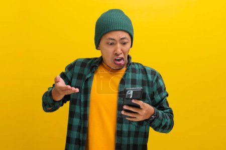 Annoyed Asian man in a beanie hat and casual shirt reacts with disdain while reading something unpleasant, possibly distasteful joke or inappropriate content on his phone against a yellow background