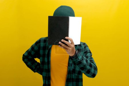 Young Asian man is covering his face with a book while standing against yellow background.