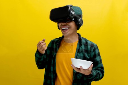 Happy Asian man wearing a VR headset munches on popcorn, seemingly enjoying a virtual movie experience. Isolated on a yellow background.