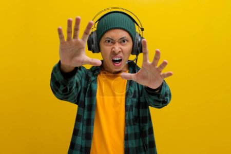 Annoyed Asian man in a beanie and casual clothes holds his hand up in a stop gesture (palm facing camera) while listening to music on headphones. Isolated on a yellow background.