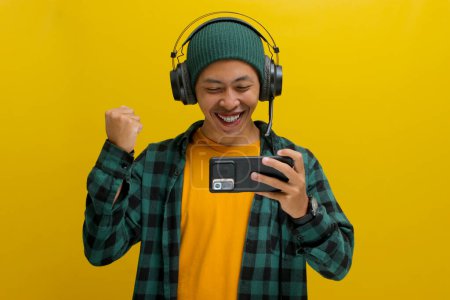 Excited Asian man in a beanie and casual clothes, wearing headphones, pumps his fist in a celebratory gesture after winning a mobile game on his phone. Isolated on a yellow background.