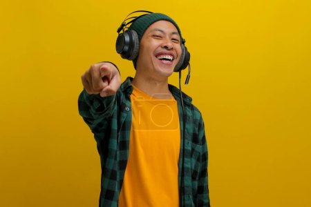 Homme asiatique excité dans un bonnet et vêtements décontractés pompe son poing vers la caméra, stimulé par la musique jouée sur son casque. Isolé sur fond jaune.