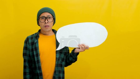 Ein betrübter junger asiatischer Mann, bekleidet mit Mütze und lässigem Hemd, hält eine Sprechblase mit einem leeren leeren Kopierraum. Sein Gesichtsausdruck zeigt Überraschung und Verwirrung, isoliert auf gelbem Hintergrund
