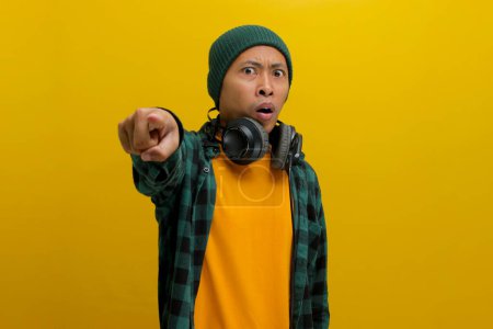 Photo for Frowning Asian man in a beanie and casual clothes, wearing headphones, points his finger in surprise towards the camera after hearing something shocking on his music. Isolated on a yellow background. - Royalty Free Image