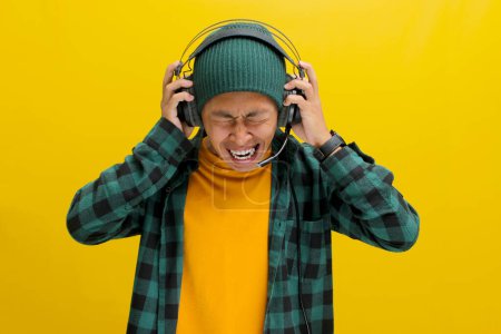 Genervter asiatischer Mann in Mütze und lässiger Kleidung ächzt vor Frustration, während er Kopfhörer trägt. Laute Musik oder Lärm werden impliziert. Vereinzelt auf gelbem Hintergrund.