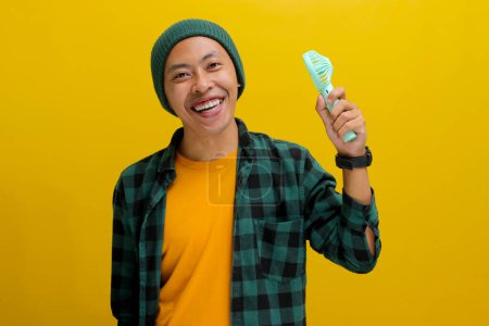 A young Asian man, dressed in a beanie hat and casual shirt, holds a handheld portable electric mini fan, relishing the cool breeze blowing against him while standing against a yellow background