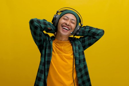 Asian man in a beanie and casual clothes stretches his muscles after work, listening to music on his headphones. A look of focus is on his face. Isolated on a yellow background.