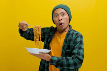Asiatischer Mann in Mütze und lässiger Kleidung macht einen überraschten und entzückten Gesichtsausdruck, während er mit der Gabel Instant-Nudeln schlürft. Vereinzelt auf gelbem Hintergrund.